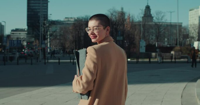 Young happy short haired business woman in beige coat, eyeglasses walks outside smiling back at camera slow motion.
