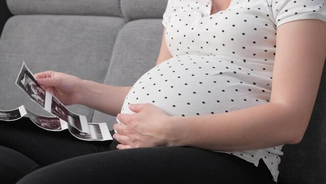 Pregnant woman looking at ultrasound photo while sitting on sofa at home. Unrecognized pregnant female holds picture with ultrasound scan and tenderly touchingy her belly.