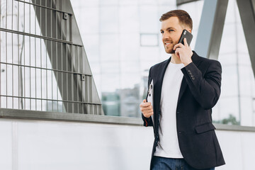 Portrait of happy modern male businessman in suit holding folder with documents and talking by phone against the background of urban buildings and offices.