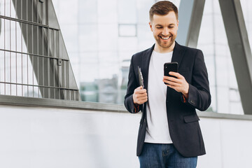 Portrait of happy modern male businessman in suit walking smiling and reading or writing message on phone on background of urban buildings and offices.