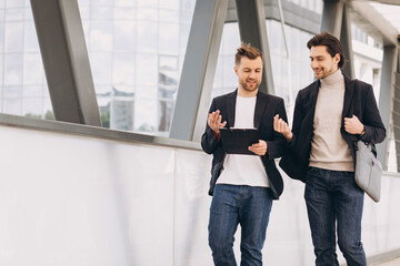 Two modern happy businessmen walking across a city bridge discussing something against the background of urban offices and buildings