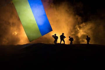 Russian war in Ukraine concept. Silhouette of armed soldiers against Ukrainian flag and burned out...