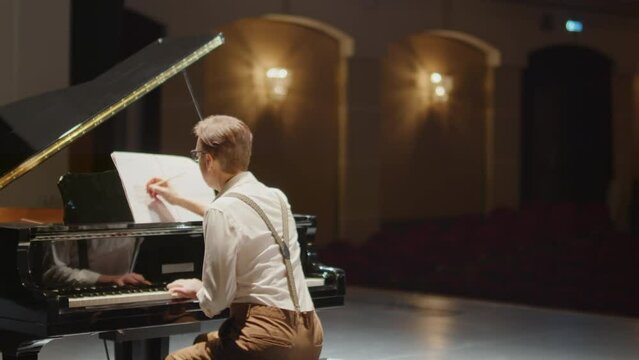 Back view of pianist composing music and writing it down on notation paper at grand piano on stage of concert hall