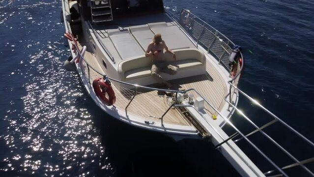 Male caucasian topless tourist resting on the deck of yacht and using smartphone in his hand filmed by drone. Aerial view of a man sunbathing on a luxury nautical vessel and surfing Internet on mobile
