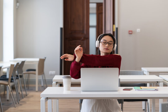 Asian girl stretching hands while watching online webinar for education distant courses or university on laptop in modern coworking space. Happy young student female has rest from study exercising