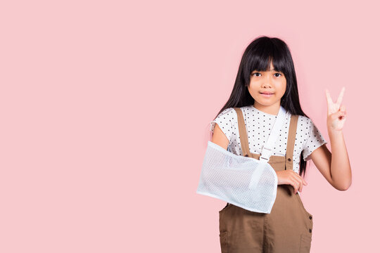 Arm broken. Asian little kid 10 years old hand bone broken from accident with arm splint show V sign at studio shot isolated on pink background, Happy child girl accident insurance and extreme sports