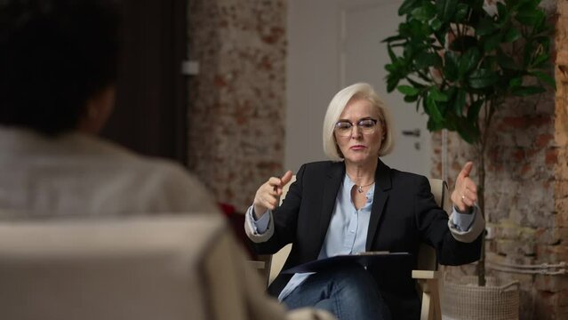 Experienced psychoanalyst is talking to female patient and sitting in modern office spbas. 4k Beautiful elderly woman talks to client and gestures with hands, looks with smile and sits in chair in