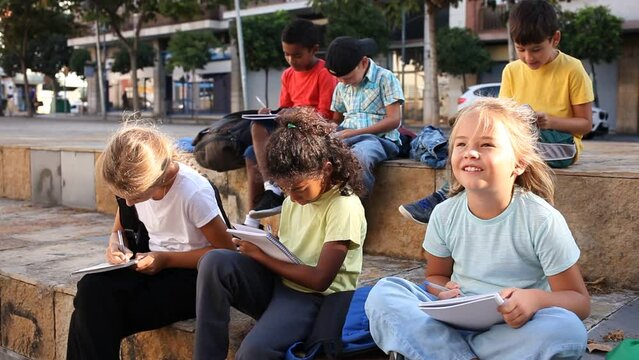 Smiling kids sitting on bench and writing in notepads, studying outdoors 