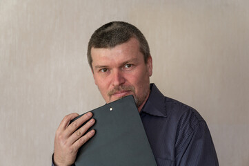 Portrait of a grown man holding a clipboard in front of his chest. Man with short haircut and graying hair looking into camera. Front view. Indoors.