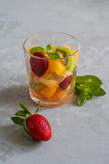 A glass with fruit salad and a mint leaf, near a ripe strawberry. Close-up.
