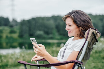 young woman freelancer sitting on chair and using smart phone. Relaxing in nature. Remote work and outdoor activity in summer.