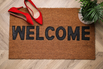 Stylish door mat with word Welcome, houseplant and shoes on wooden floor, top view