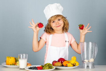 Child chef cook hold strawberries prepares food on isolated grey studio background. Kids cooking. Teen boy with apron and chef hat preparing a healthy vegetables meal in the kitchen.