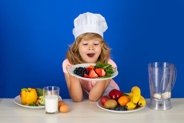 Child wearing cooker uniform and chef hat hold plate with fruits preparing vegetables on kitchen, studio portrait. Cooking, culinary and kids food concept.