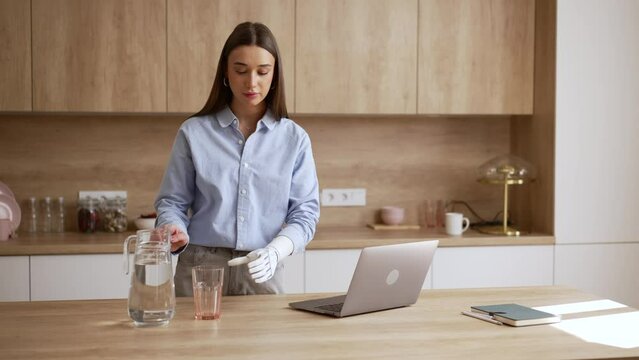 Young woman is using laptop and drinking water at table in home kitchen spbas. 4k Beautiful American female looks at display and surfs web or works then pours fresh water into glass and drinks with