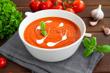 Tomato soup puree in a bowl with cream and fresh basil leaves on a dark wooden background. Copy space. Top view.