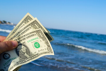 Person holds in his hand three dollar bills on the background of the waves of the blue sea. Concept money finance business tourism travel, money spending, cash expenses. Money cash expenses business - obrazy, fototapety, plakaty