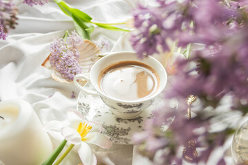 Fototapeta na wymiar Spring composition with tulips and lilac flowers, candles and cup of coffee on white bed sheets. Cozy home atmosphere.