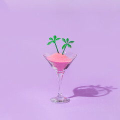 Summer tropical creative layout with martini cockatil glass with pink sand and palm trees figurines on pastel purple background. 80s, 90s retro fashion aesthetic summer concept. Minimal drink concept.