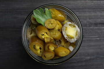 Glass jar with slices of pickled green jalapeno peppers on black wooden table, top view