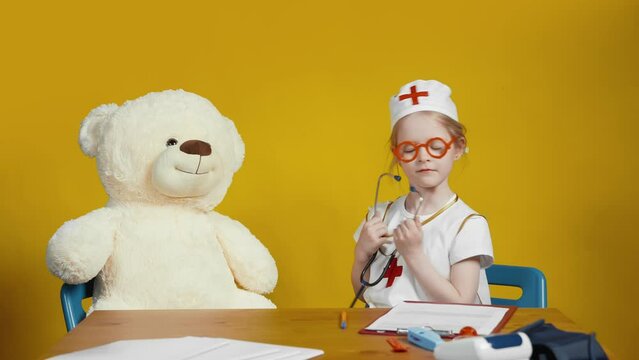 A little girl veterinarian in a white suit listens to the heartbeat of a teddy bear with a stethoscope on a yellow background. A little girl plays a doctor. Children as adults