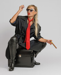 young blonde woman with long hair in sunglasses with a cigar in her hand sits on a suitcase on a...