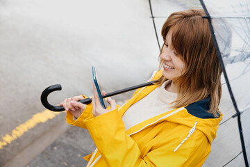 young caucasian woman dressed in a yellow raincoat holds a transparent umbrella and uses a...