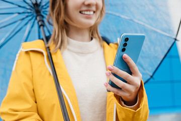 young caucasian woman dressed in a yellow raincoat holds a transparent umbrella and uses a smartphone, close-up