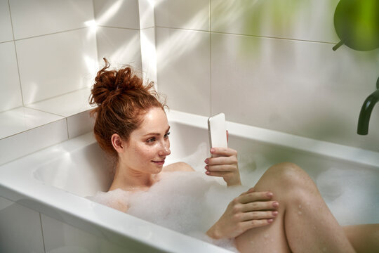 Caucasian redhead woman taking a bath and using mobile phone - Powered by Adobe