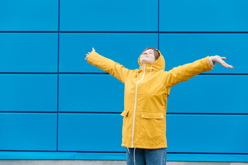 A young woman in a yellow raincoat stands with her arms outstretched against a blue wall