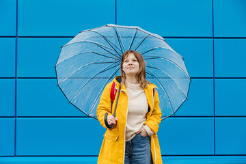 portrait of a pretty girl dressed in yellow raincoat posing while standing with an open umbrella over blue background