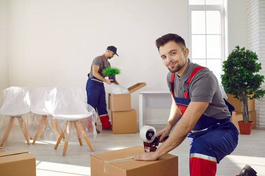 Portrait of happy young man in uniform looking at camera while packing and sealing box using sticky adhesive duct tape gun roller dispenser. Moving house, relocation company, delivery service concept