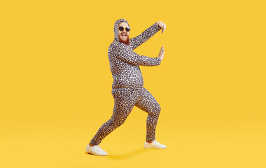Fototapeta na wymiar Funny chubby guy in crazy costume having fun in modern fashion studio. Happy cheerful confident fat man wearing comfortable leopard pajamas dancing isolated on yellow background. Crazy party concept
