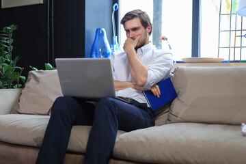 Fototapeta na wymiar Unhappy frustrated young male holding head by hand sitting with laptop on sofa.