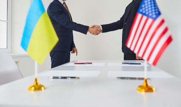 Male politicians from America and Ukraine shake hands after agreeing to sign lend lease for Ukraine. Flags of countries in blurred foreground against background of handshake of diplomats.Cropped image - Powered by Adobe