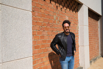 Fototapeta na wymiar middle-aged man, handsome and dark, bearded and with a sculpted body on a brick wall. Man wearing black leather jacket, mirrored sunglasses, and jeans. Man poses for photo. Men's fashion concept.