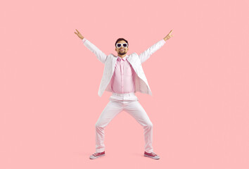 Full length photo of a young fashionista dancing in the studio. Happy handsome young guy in a white suit, pink shirt, pink sneakers and sunglasses raised his hands in the air on a pink background