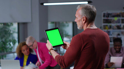 Back view of mature man holding digital tablet with green screen standing in modern office