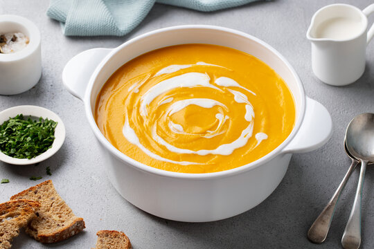 Pumpkin, vegetable cream soup in white pot, saucepan, bowl. Grey background. Close up.
