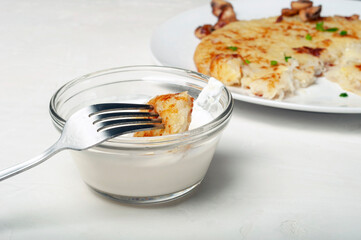 on fork is a piece of potato tortilla - a traditional Swiss dish, the fork is raised over a saucepan with cream sauce. in the background, you can see part of a plate with a potato pancake, side view.