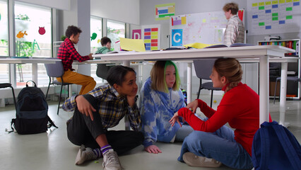 Happy diverse teen girlfriends sit under desk in class and gossip during break