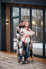 Full length of man wrapped in blanket kissing girlfriend in cheek while woman holding bouquet of flowers. Happy romantic couple standing outside scandinavian house barnhouse under winter snow.
