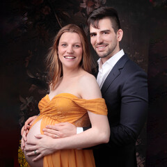 Beautiful young pregnant woman wearing a maternity chiffon pregnancy dress with a handsome young man wearing a suit with flowers in the background. 