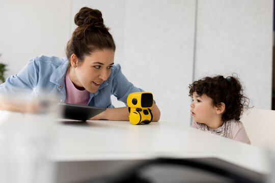 Working mother sitting at table daughter playing with AI toy robot