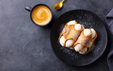 Cannoli Italian dessert on a black plate with cup of coffee. Grey background. Copy space. Top view.