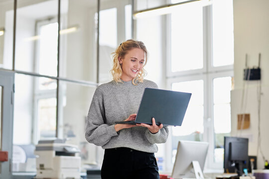 Happy young woman standing in office using laptop