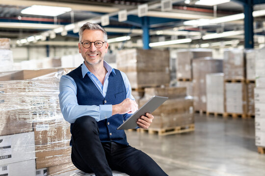 Happy manager wearing eyeglasses with tablet PC sitting in front of cardboard box in warehouse