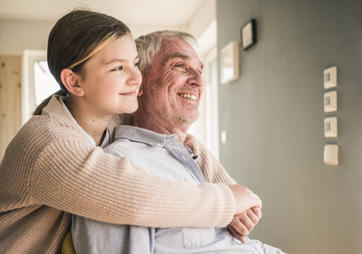 Smiling girl hugging grandfather from behind at home