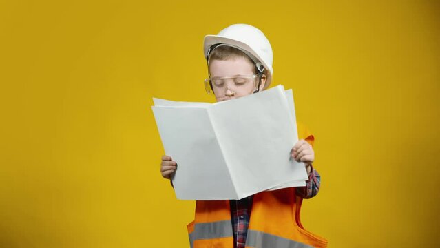 Portrait of a little construction boy in a helmet, looking at building diagrams on paper, standing on a yellow background. Children as adults