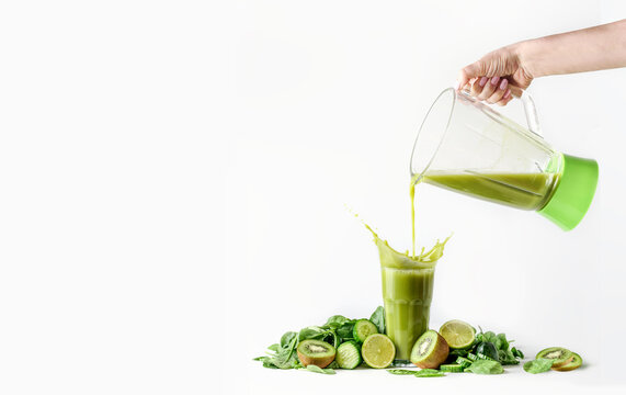 Women hand pouring green smoothie from blender in glass with splashing and ingredients: kiwi, cucumber, lime and spinach at white background. Healthy lifestyle, dieting and detox food